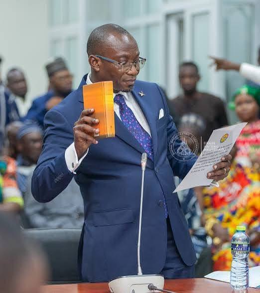 A man in a navy suit and glasses holding a booklet and a paper at a microphone